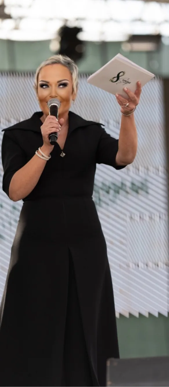 Sahne: Özlem uzun siyah elbiseyle mikrofonla konuşuyor, elinde kart; arkada etkinlik panosu.