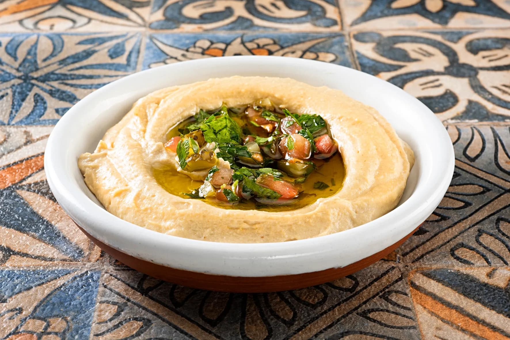 Beyaz kâsede humus, ortasında zeytinyağı, domates ve maydanozlu salata; desenli çini zemin. Yatay.