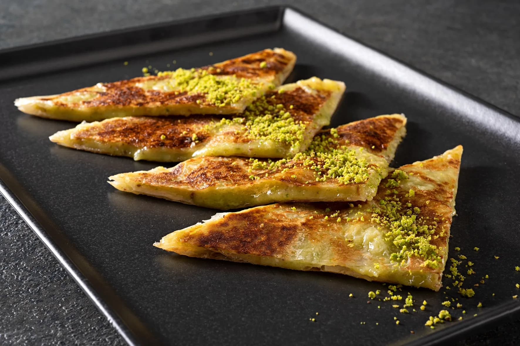 Siyah tepside üçgen kesilmiş ince börek dilimleri, üstünde yeşil fıstık. Yatay.