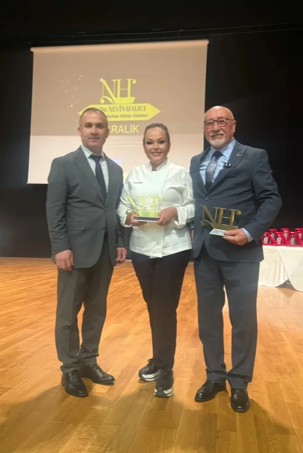 Tören sahnesi: Özlem beyaz şef ceketiyle ortada, elinde altın renkli 'NH' ödül heykelciği; iki yanında takım elbiseli iki erkek; arkada perdede 'IV. Dr. Nevin Halıcı' logosu. Dikey.