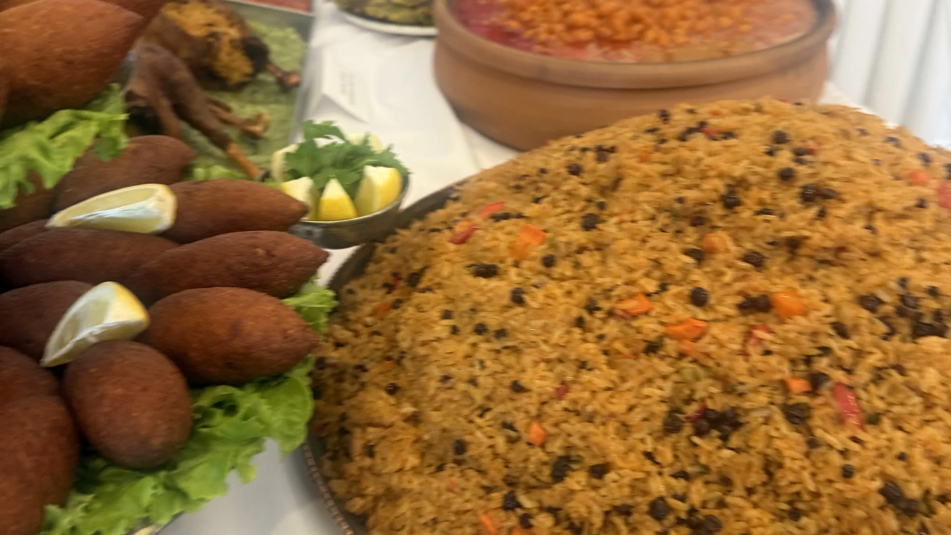 Yakın çekimde bir sofra: büyük tepside kuş üzümlü ve biberli pilav, yanında marul yaprakları üzerine dizilmiş kızarmış köfteler ve limon dilimleri, arkada toprak kapta kırmızı sulu bir yemek