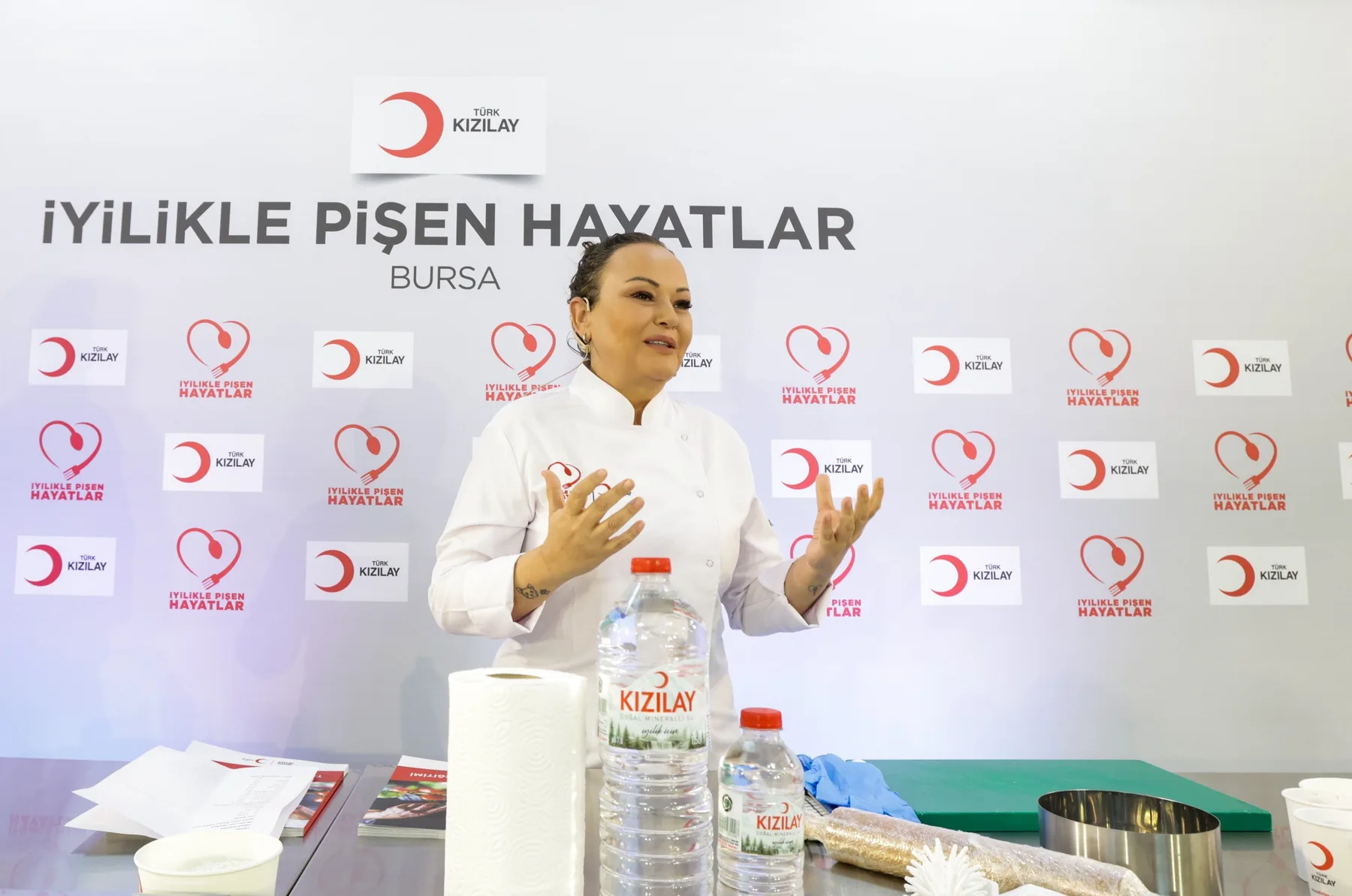 Türk Kızılay · İyilikle Pişen Hayatlar, Bursa: Özlem beyaz şef ceketiyle tezgâhın arkasında iki elini açmış anlatıyor; arkada Kızılay panosu, önde su şişeleri, kâğıt havlu, yeşil kesme tahtası.