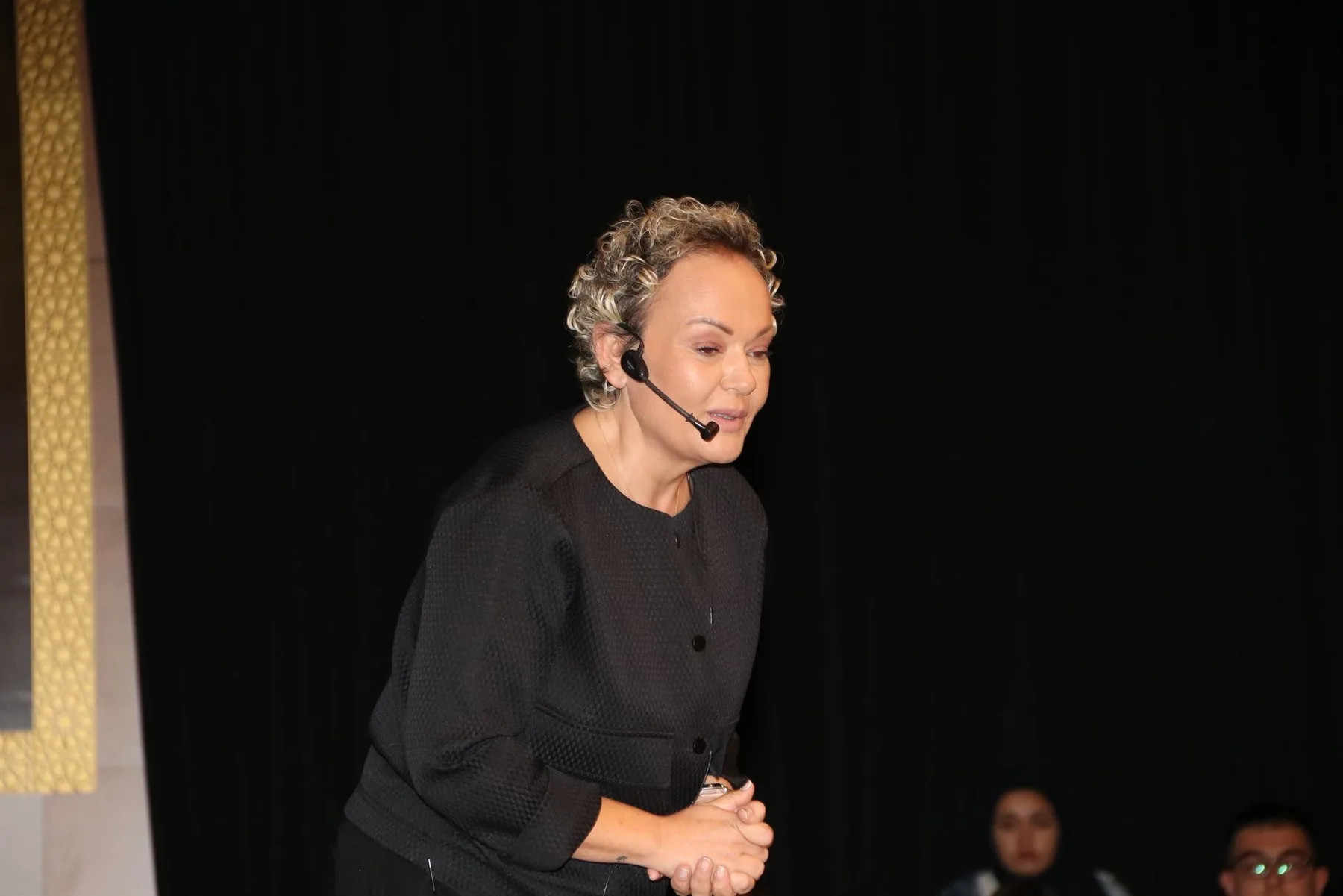 Aynı salon: Özlem öne eğilmiş, ellerini birleştirmiş konuşuyor; önde seyirci.