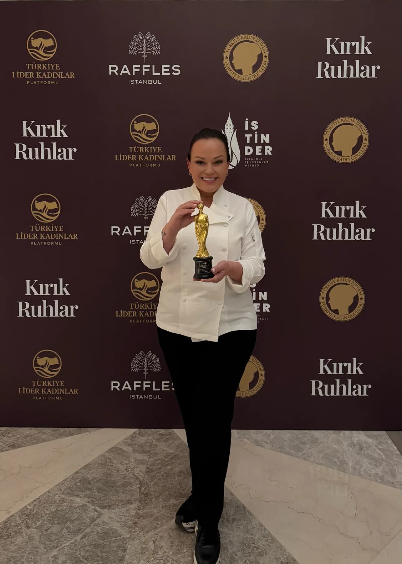 2026 ödülü: Özlem beyaz şef ceketiyle bir basın panosunun önünde, elinde altın heykelcik, gülümsüyor; panoda Raffles İstanbul, İSTİNDER, Türkiye Lider Kadınlar Platformu ve 'Kırık Ruhlar' yazıları. Dikey.