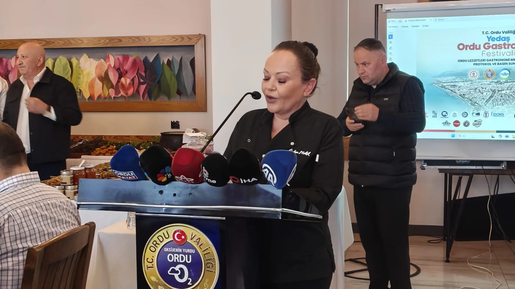 Ordu Gastronomi Festivali basın toplantısı: Özlem siyah şef ceketiyle kürsüde konuşuyor, kürsüde televizyon kanallarının mikrofonları, iki yanında iki erkek protokol üyesi, ekranda festival afişi.