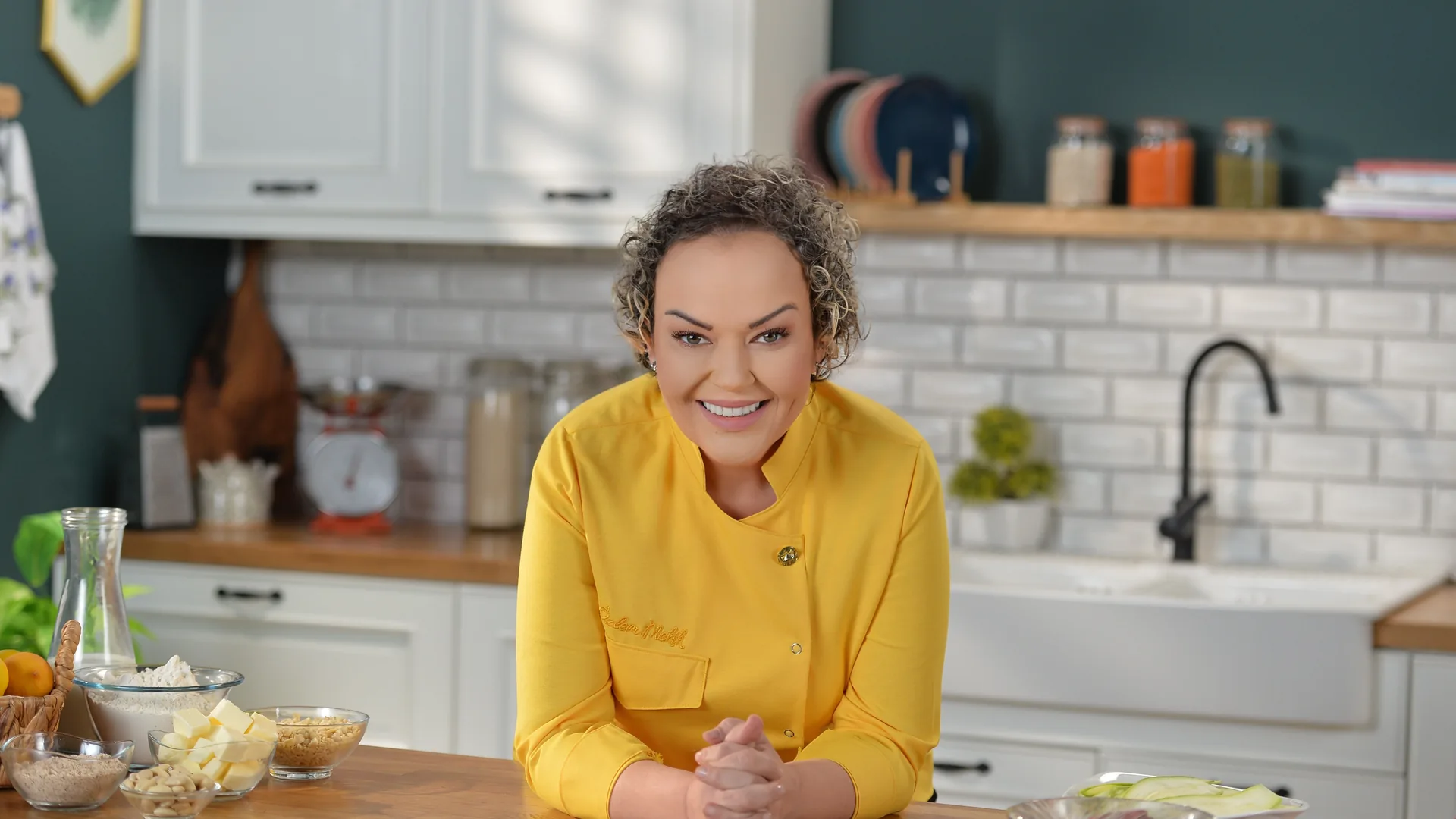 Özlem Mekik in a yellow chef's jacket behind a wooden kitchen counter, hands clasped, smiling; glass bowls of flour, butter and nuts on the counter and spice jars on the shelves behind her