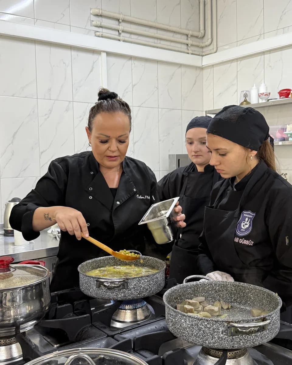 Özlem Mekik in a black chef's jacket stirring a pan on the stove with a wooden spoon while a participant in an apron passes her an ingredient