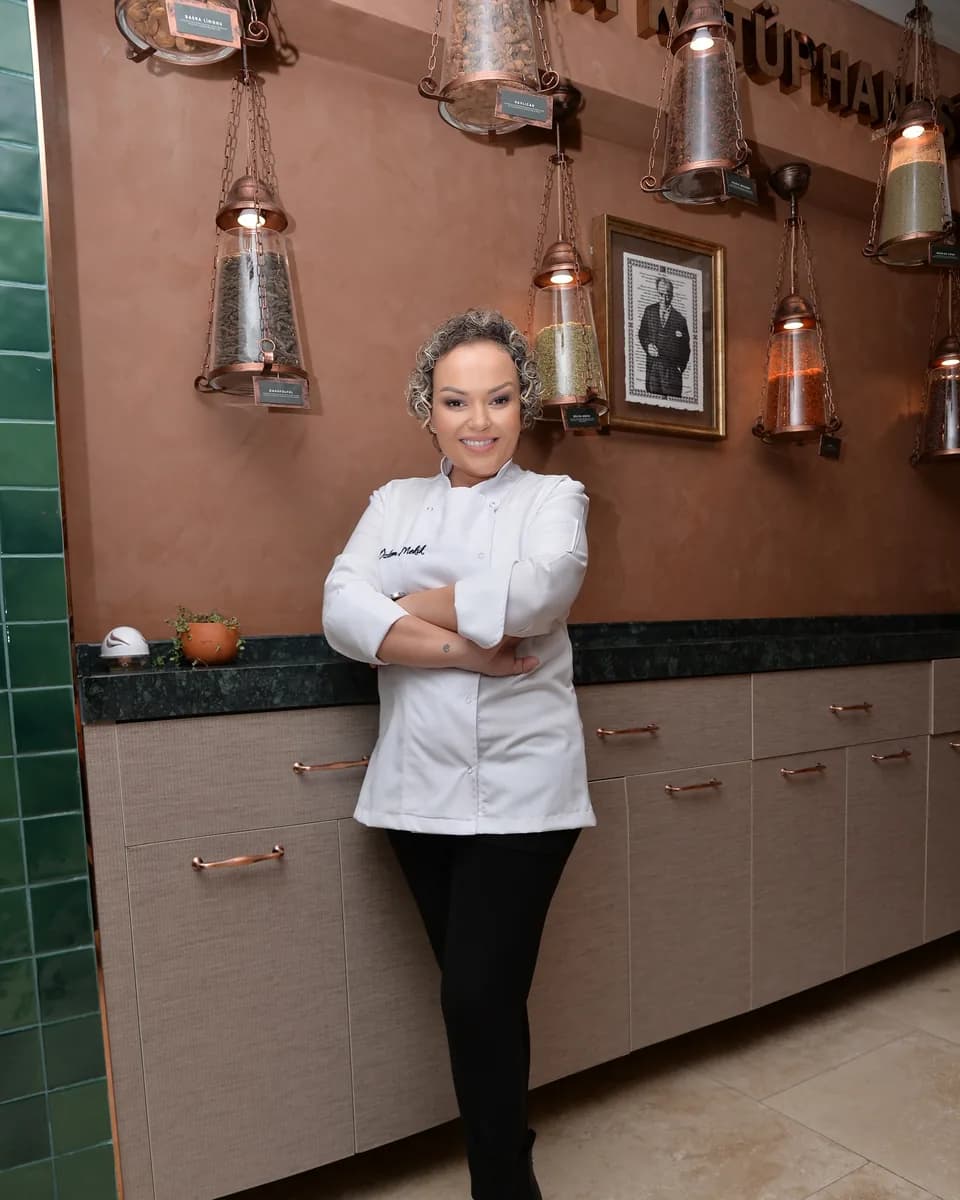 Özlem Mekik in a white chef's jacket leaning against wooden cabinets with her arms folded; glass and copper storage jars hang on the wall behind her