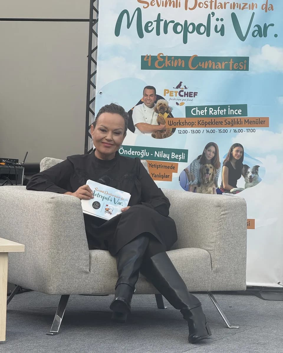 Özlem Mekik seated in a grey armchair holding a board on her lap, with a large event poster on the wall behind her